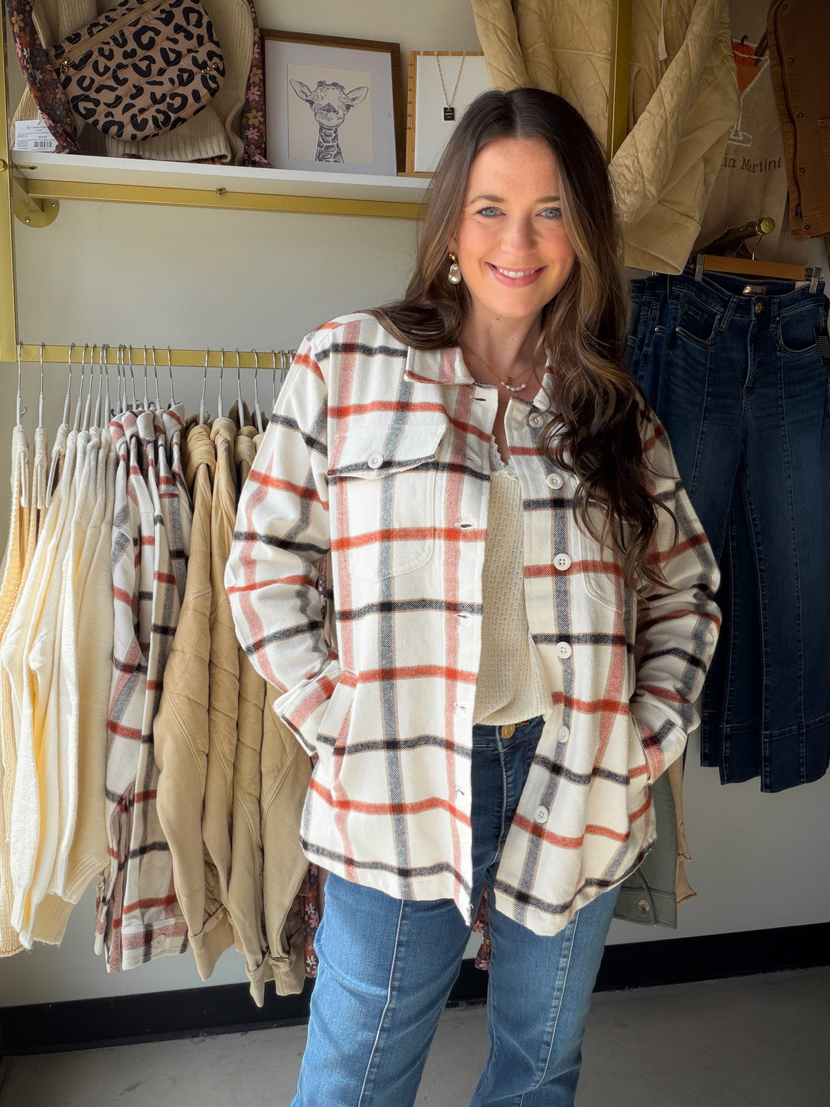 Model wearing an ivory and rust plaid shacket with a button front and chest pockets over a cream top and jeans.