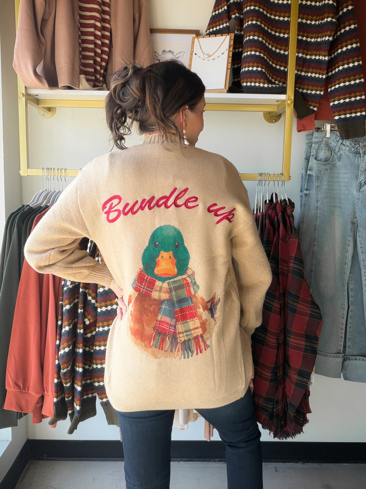A Cozy Layer with a Playful Surprise

Classic from the front, full of personality from the back. The Bundle Up Sweater Cardigan by Anniewear brings a charming twist to your cool-weather wardrobe with a scarf-wearing duck and cheerful red “Bundle up” lettering across the back. Its warm beige color keeps the look easy to pair, while the colorful plaid scarf adds just the right touch of seasonal fun.

Slip it on for a coffee run, a weekend gathering, or an afternoon exploring your favorite little shops. The op