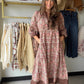 A woman modeling a long-sleeve, knee-length tunic dress with a pink, brown, and beige bohemian floral pattern, standing in a boutique clothing store.