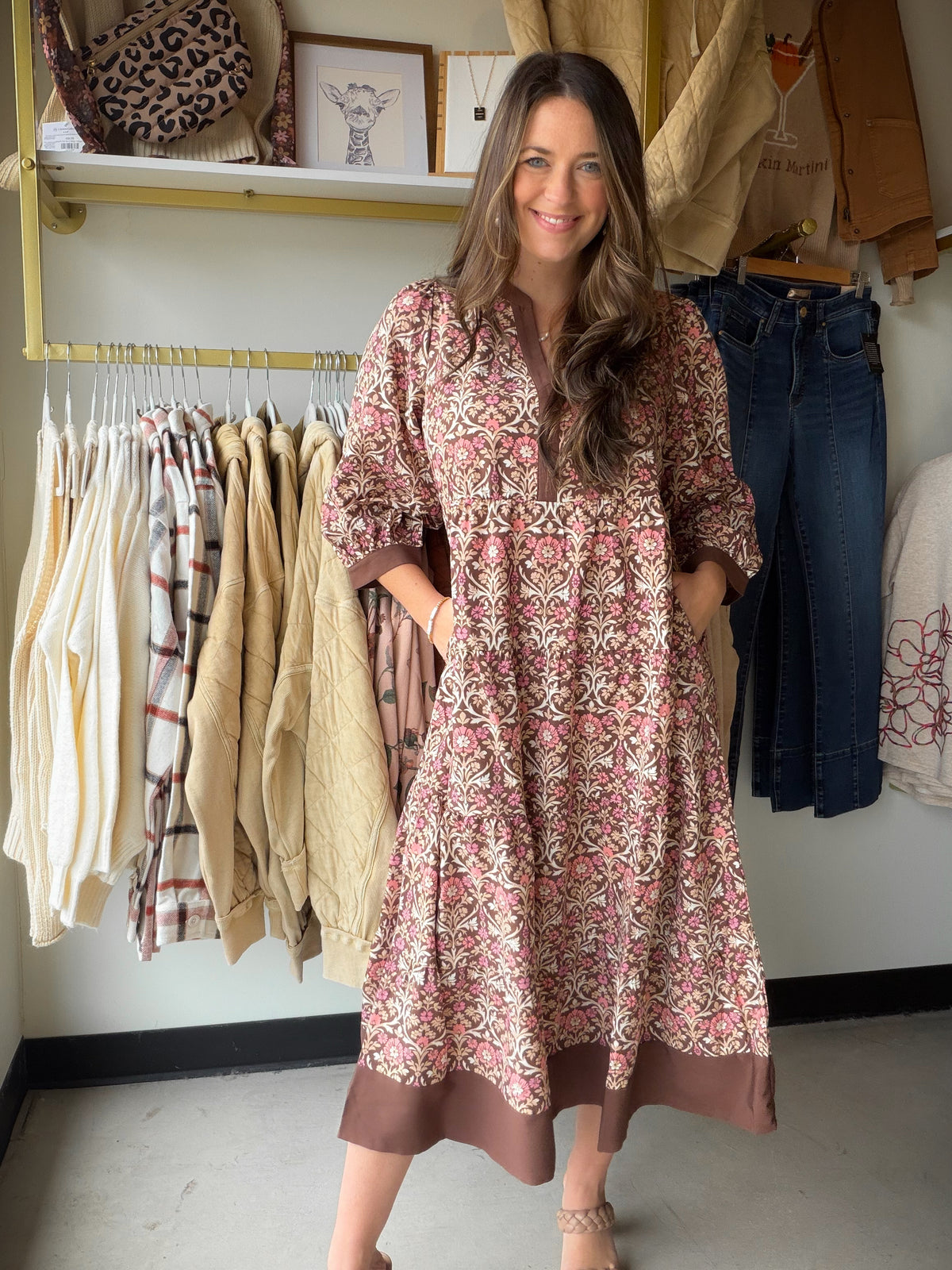 A woman modeling a long-sleeve, knee-length tunic dress with a pink, brown, and beige bohemian floral pattern, standing in a boutique clothing store.