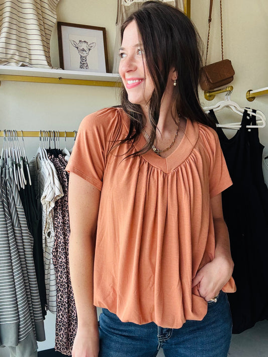 This Butter Orange Shirring Short Sleeve top features a flattering V-neck and short sleeves, designed with elegant shirring details for a refined silhouette. Its long length offers versatile styling options, making it suitable for both casual and polished looks. Crafted to combine comfort with a sophisticated touch, this top is an excellent addition to any wardrobe seeking effortless style.